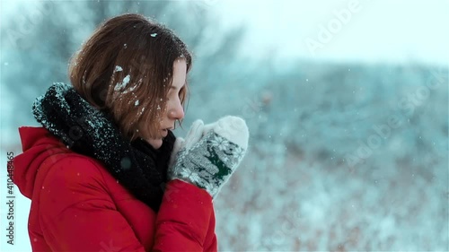 A gentle caucasian girl is standing in the snow and with a slight smile on her face warms her hands in the snowfall, blows mouth on her gloves  and snowflakes stick to her brown hair. Side view