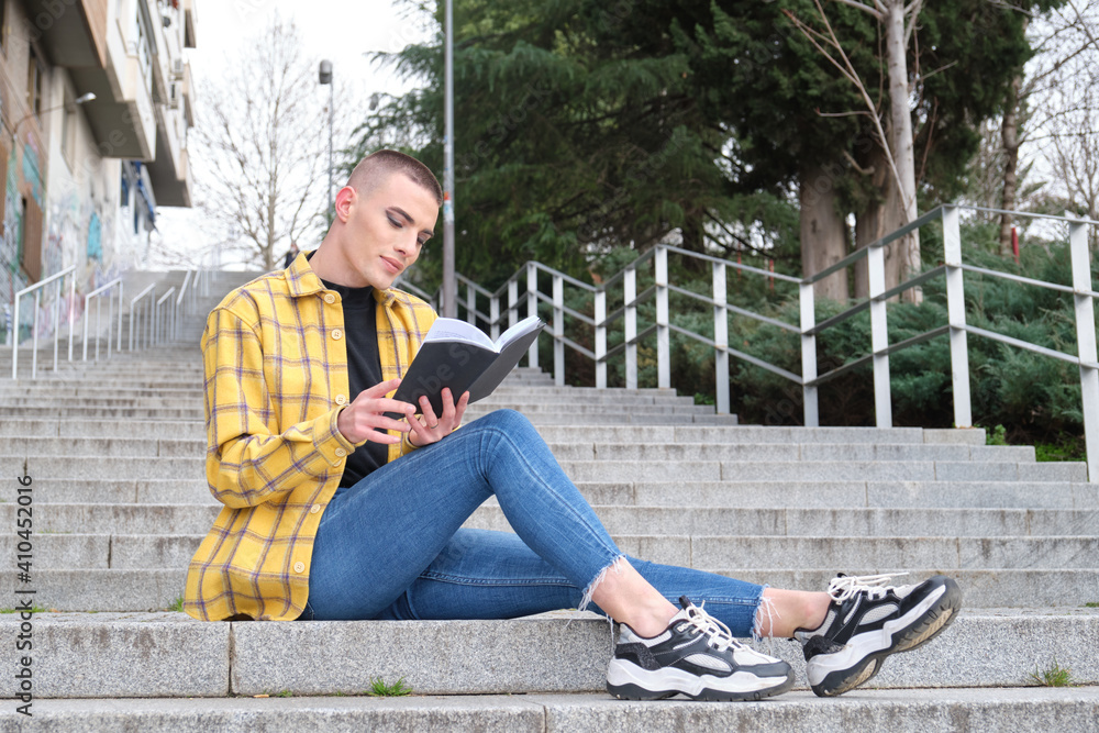 Handsome young man wearing make up, smiling and reading a book sitting on stairs. Non binary androgynous guy. Handsome young man wearing make up, smiling and reading a book sitting on stairs. Non binary androgynous guy.