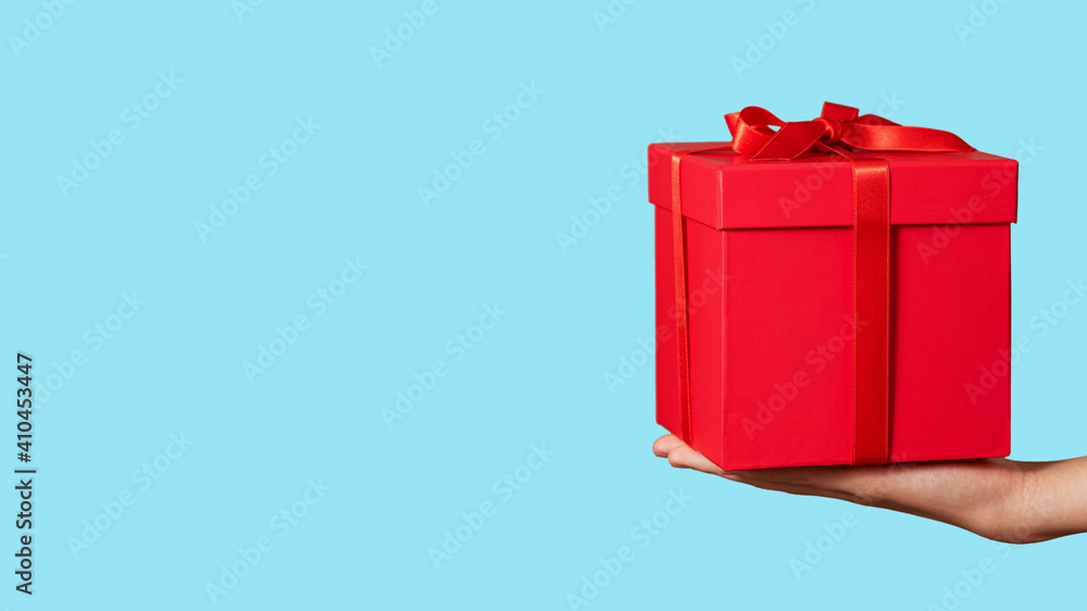 Hand holding red gift box with ribbon bow isolated on light blue background. Holiday concept. Copy space.