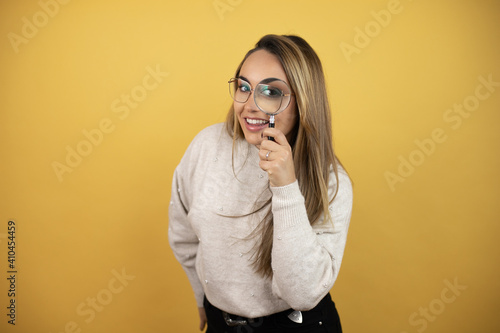 Pretty woman with long hair looking through a magnifying glass against yellow wall