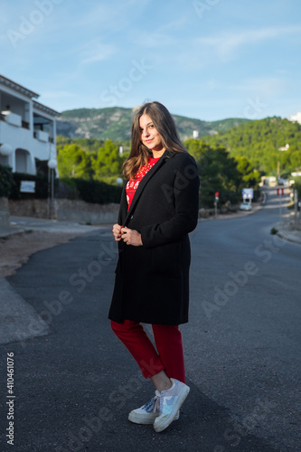 Beautiful young blond woman wearing a long black coat, red pants and white sneakers. In the street, smiling and looking at the camera. Professional vertical shot. Alcossebre, Spain. 