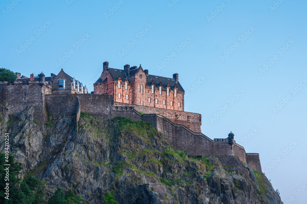 Steep cliffs with walls and the hospital building of Edinburgh castle seen from the north under a blue sky during sunset.