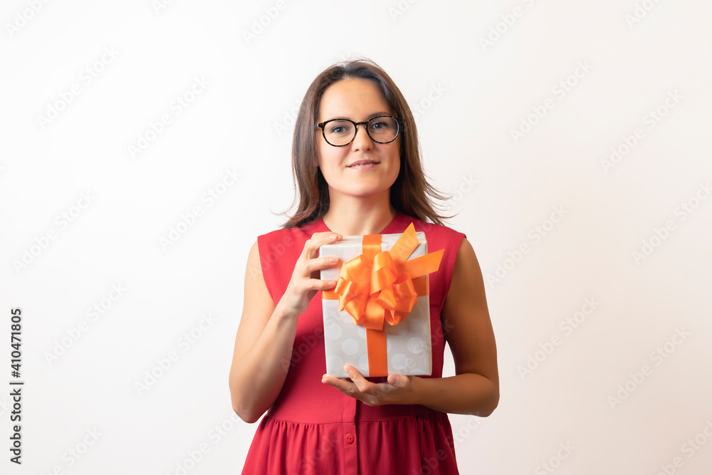 A young woman in a red dress and glasses holding a box with red ribbon, considering, twists a gift for the holiday. The girl looks at the new gift and smiles, becomes happy A young woman in a red dress and glasses holding a box with red ribbon, considering, twists a gift for the holiday. The girl looks at the new gift and smiles, becomes happy