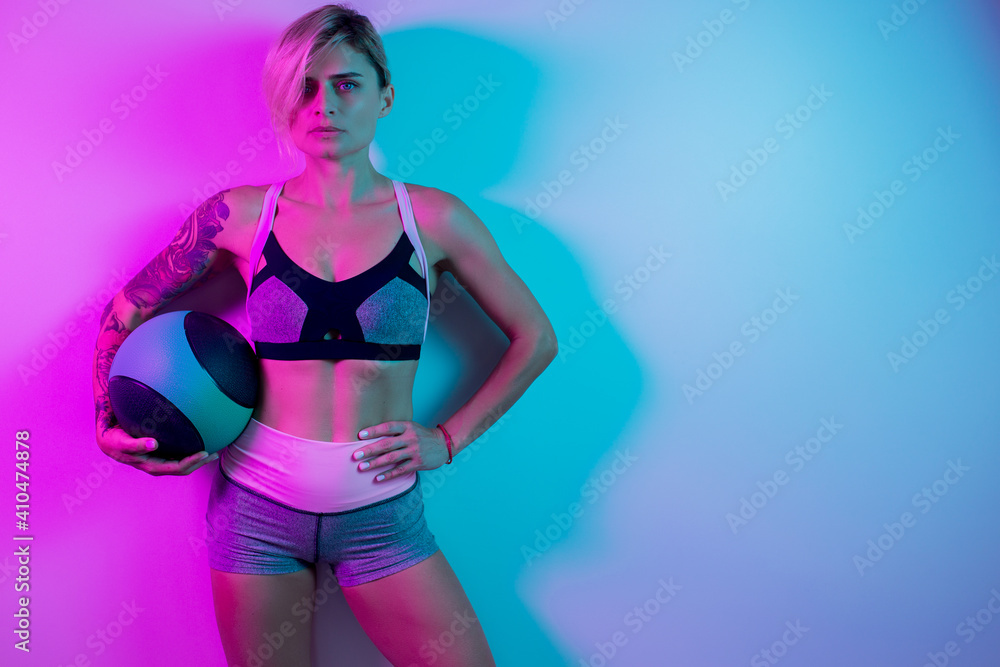 Obraz premium Fit young blonde woman posing with medicine ball in gym in neon lights.