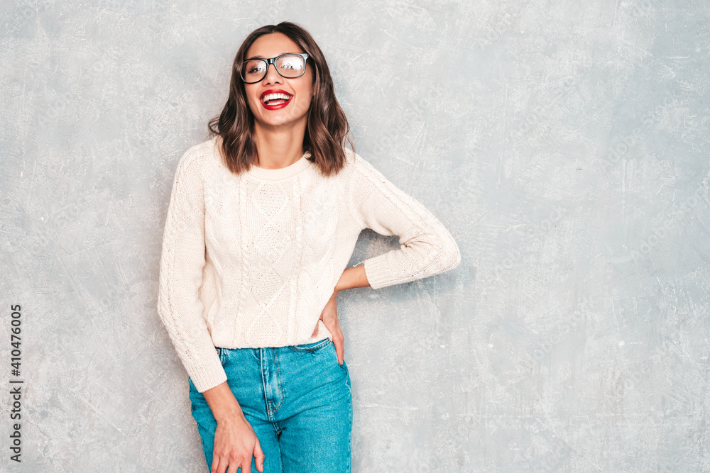 Young beautiful smiling woman. Trendy female in casual summer jeans clothes. Positive female with red lips shows facial emotions. Funny and cheerful model posing near gray wall in studio in eyewear