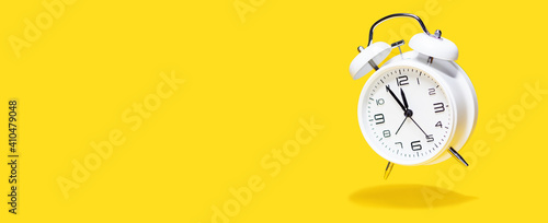 White bell alarm clock hovering over yellow background. 5 to 12 clock concept isolated