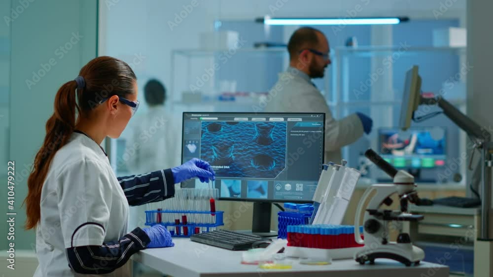 Chemistry doctor analysing blood sample and typing on computer while coworker opening door using digital computer with touch scan. Woman examining virus evolution using high tech and chemistry tools Chemistry doctor analysing blood sample and typing on computer while coworker opening door using digital computer with touch scan. Woman examining virus evolution using high tech and chemistry tools