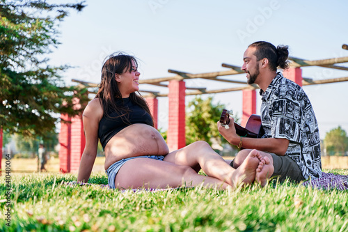 Portrait of two young and future parents in a park on a blanket and him playing the guitar and singing to her, pregnancy and new family concept