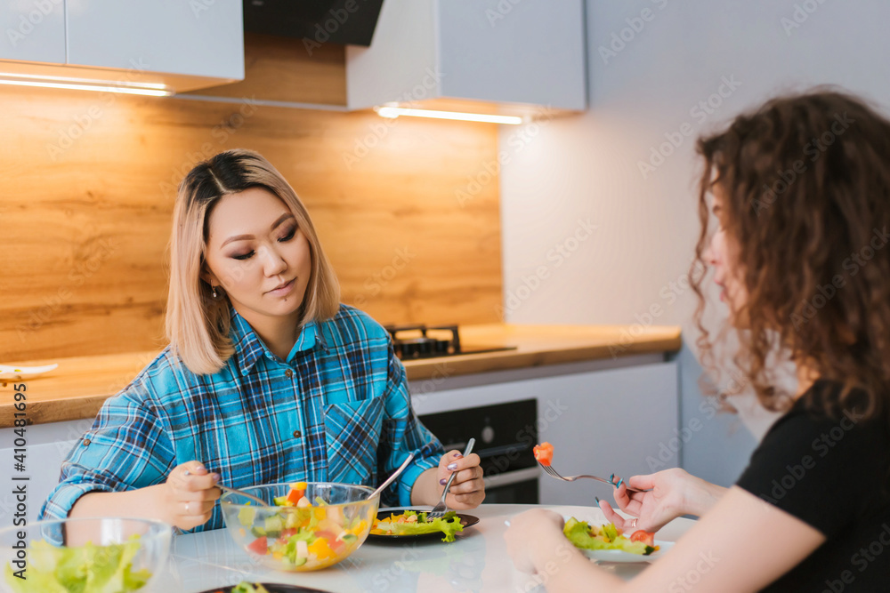 Two girls are sitting at the kitchen table and eating a diet salad. Girlfriends chatting over food. Two girls are sitting at the kitchen table and eating a diet salad. Girlfriends chatting over food.