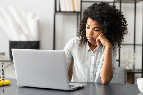 Young African American female trainee or an office worker with Afro hairstyle sitting at the desk, feeling bored and sleepy does not want to do homework or work on the project, unhappy with the task photo