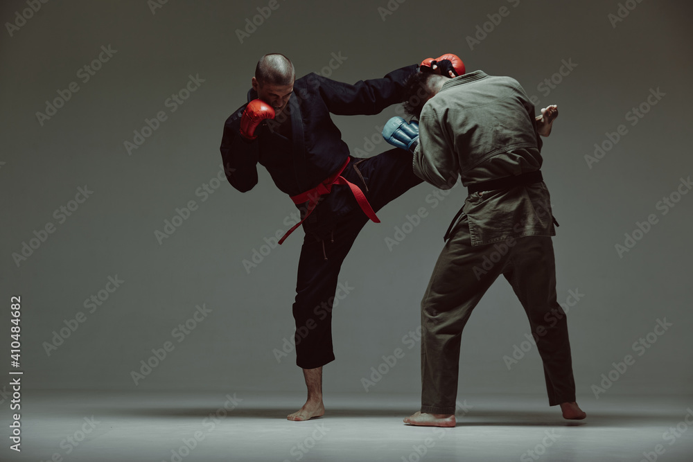Sportive males fighting, wearing kimono and boxing gloves on gray studio backdrop with copy space, mixed fight concept