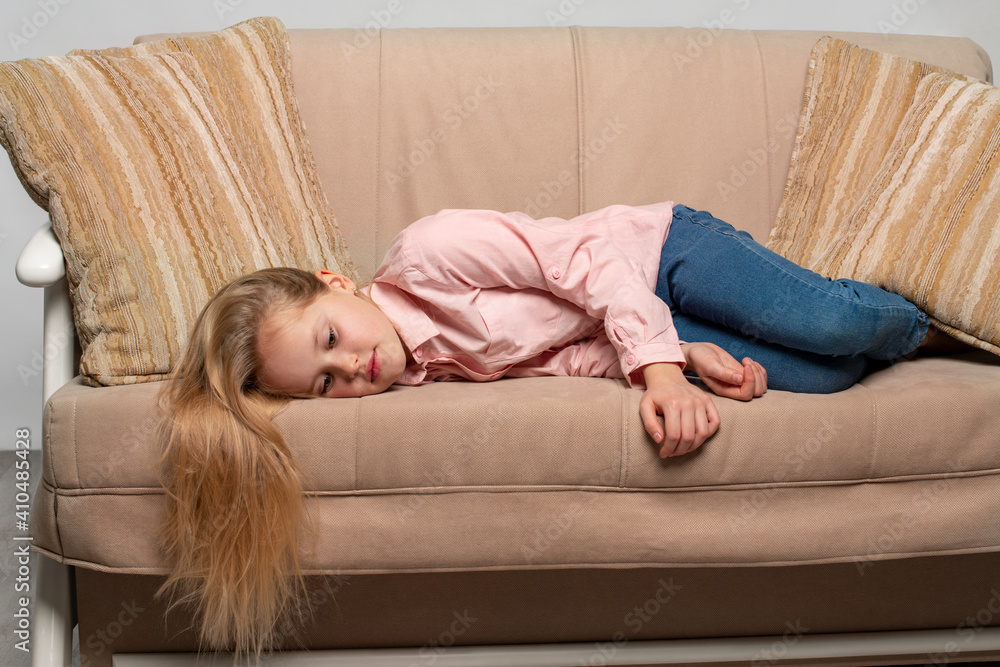 A little blonde girl with long hair wearing pink shirt and jeans lies on the couch and is sad. A little blonde girl with long hair wearing pink shirt and jeans lies on the couch and is sad.