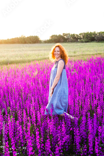 A young beautiful woman with red hair walks in a field with purple flowers in the rays of the setting sun. Active rest, nature walks, digital detox, unity with nature, alone with yourself.
