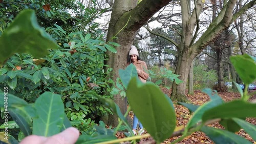 A woman on her phone in the winter time wearing a woolly jumper and pink bobble hat in a wooded park with somebody looking and spying at her through branches and bushes, voyeurism stalking concept.