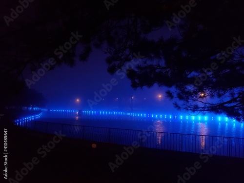 Bright blue illumination with blurry reflection in pond water. Landscape of public park with lake illuminated by blue spotlights glowing in night darkness