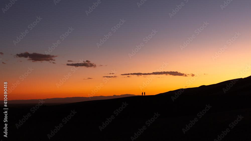 Wide colorful shot of silhouette of two people standing on top of sandy dune at orange sunset in america