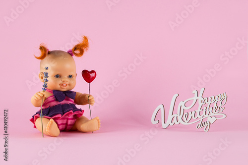 Freckled white doll girl with two curly red tails holds a heart and a sprig of lavender on a pink background. Space for inscriptions, postcard. Happy Valentine's day