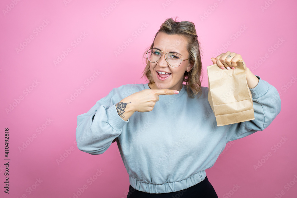 Young caucasian woman wearing sweatshirt over pink background smiling, surprised and pointing paper bag in her hand Young caucasian woman wearing sweatshirt over pink background smiling, surprised and pointing paper bag in her hand