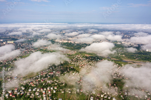 Aerial view of white clouds above a town or village with rows of buildings and curvy streets between green fields in summer. Countryside landscape from above.