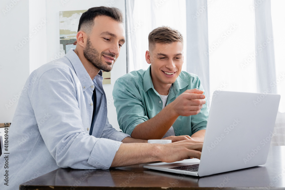 homosexual man pointing with finger and looking at laptop near happy husband homosexual man pointing with finger and looking at laptop near happy husband