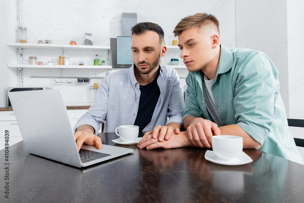 homosexual couple looking at laptop near cups on table homosexual couple looking at laptop near cups on table
