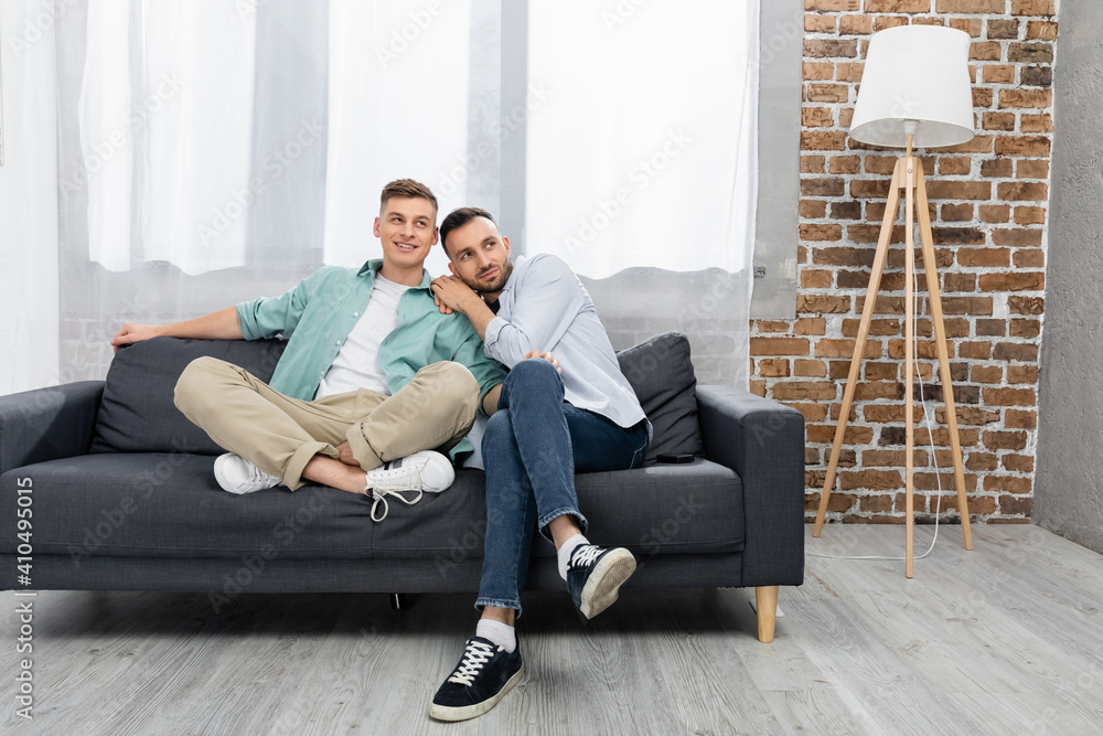 cheerful same sex couple looking away while sitting on couch at home cheerful same sex couple looking away while sitting on couch at home