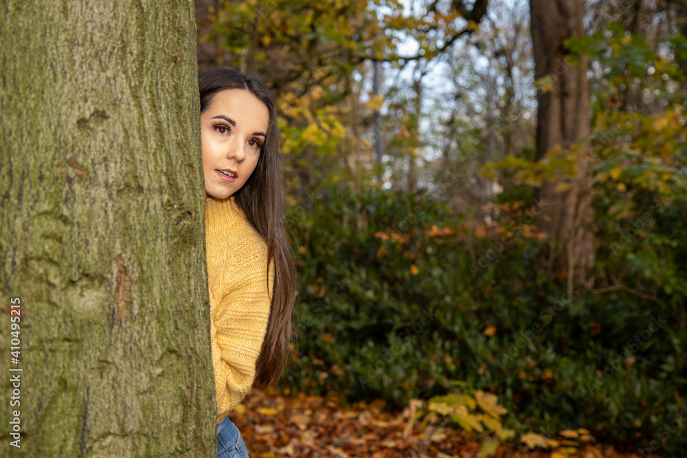 A young attractive woman with long brown hair in the winter time in a wooden park area wearing a yellow jumper looking out from behind a tree. A young attractive woman with long brown hair in the winter time in a wooden park area wearing a yellow jumper looking out from behind a tree.