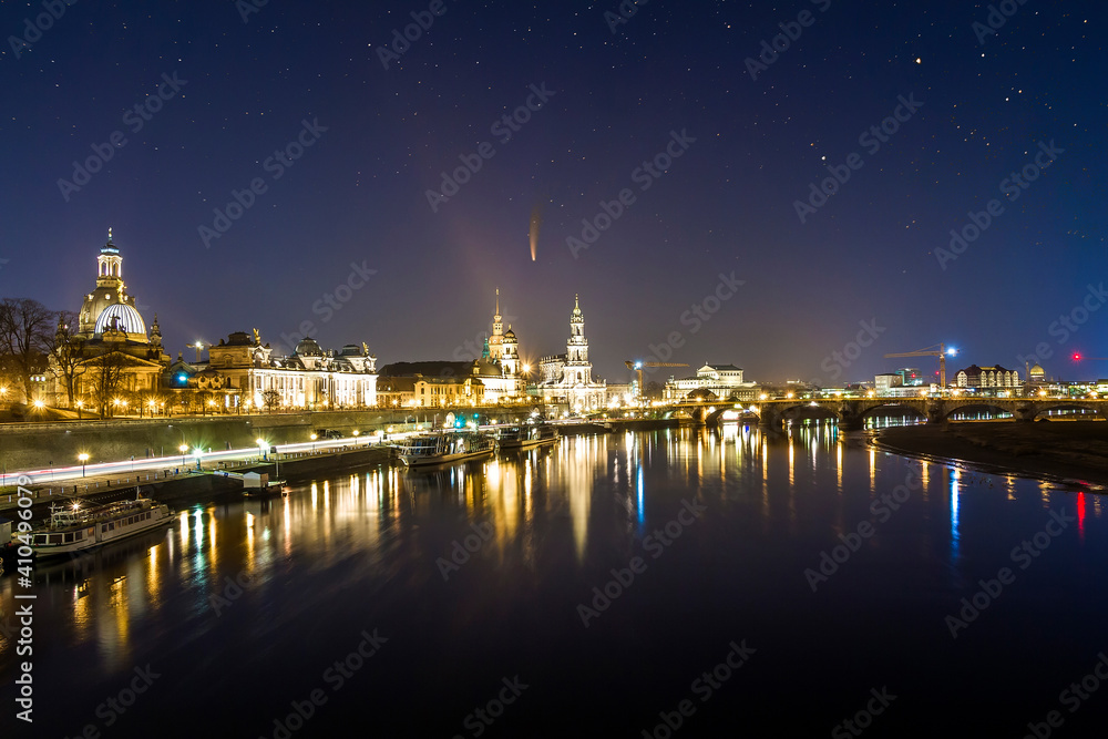 Night cityscape view of historic buildings with reflections in Elbe river in the center of Dresden (Germany) and starry dark sky with Neowise comet with light tail.