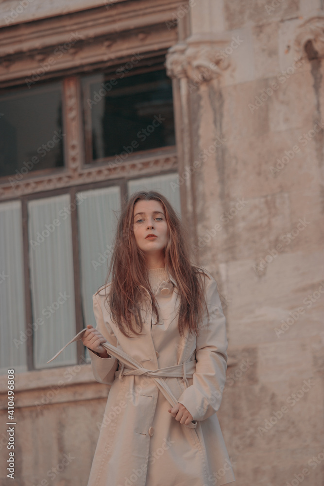 Portrait of an elegant young woman outdoors with a casual attire and brown jacket posing and looking at the camera in city center of a brown wall background while tying jacket.