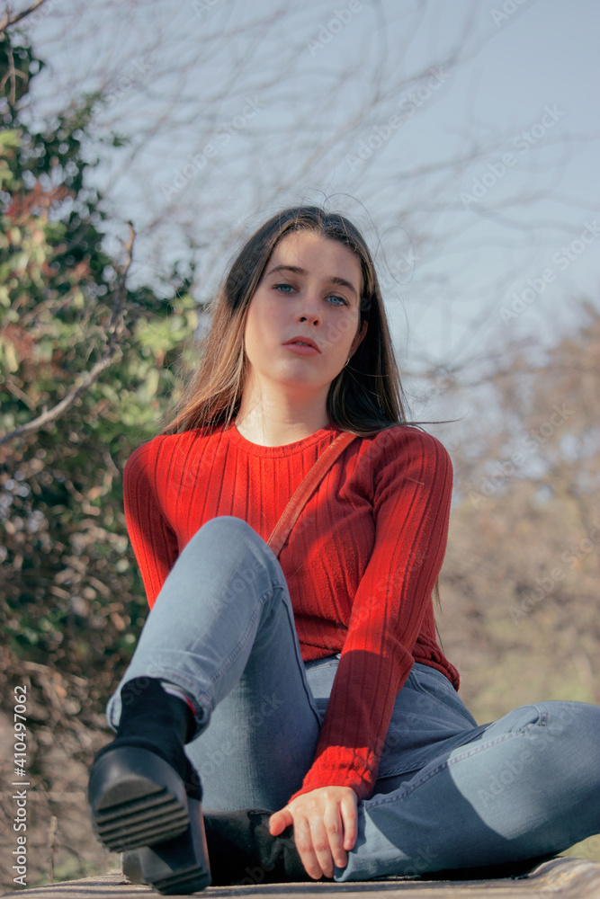 Obraz premium Close up picture of young thoughtful woman in red jersey and black high heels looking in front at the camera in a winter sunny day while sitting on a bench in side of a park.