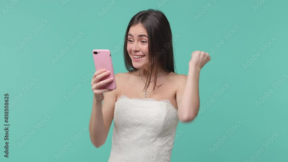 Surprised joyful bride young woman in white wedding dress posing isolated on blue turquoise background studio. Wedding concept. Using mobile phone typing browsing say wow yes doing winner gesture Surprised joyful bride young woman in white wedding dress posing isolated on blue turquoise background studio. Wedding concept. Using mobile phone typing browsing say wow yes doing winner gesture
