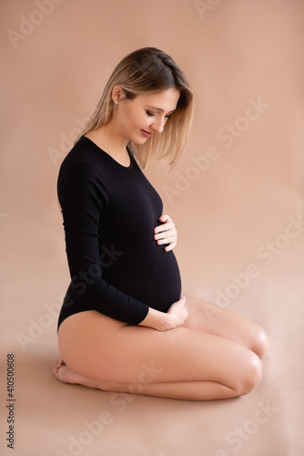 Happy pregnant woman 25-29 year old wearing black stylish body suit sitting on floor over beige background close up. Motherhood. Materity. photo