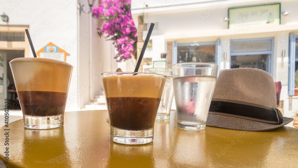 two ice coffees and two glasses of water and a hat in focus on a table with a blurred background of sunny white houses on Kythira, Greece two ice coffees and two glasses of water and a hat in focus on a table with a blurred background of sunny white houses on Kythira, Greece