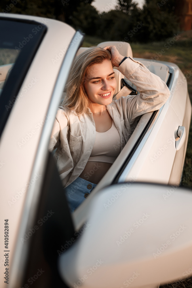A glamorous luxury girl with blonde hair smiling while sitting on a white convertible. Young successful woman sitting in her white car A glamorous luxury girl with blonde hair smiling while sitting on a white convertible. Young successful woman sitting in her white car