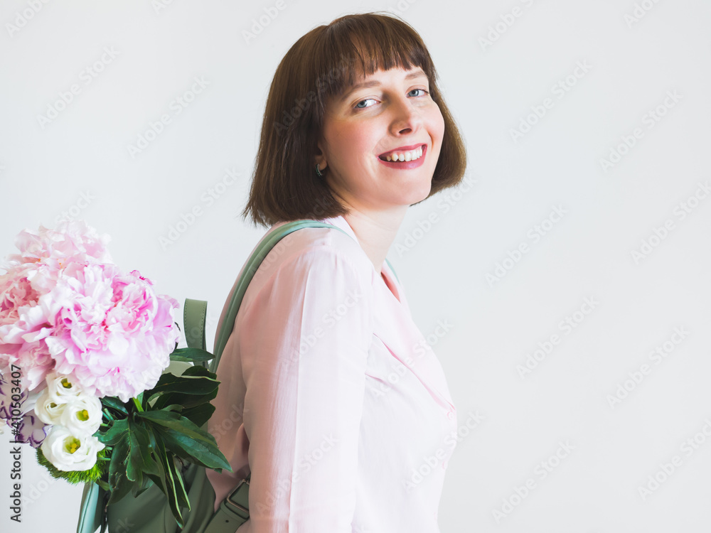 Young woman wearing pink shirt and backpack with pastel peony bouquet on gray wall background. Lifestyle spring concept. Mothers day gift Young woman wearing pink shirt and backpack with pastel peony bouquet on gray wall background. Lifestyle spring concept. Mothers day gift