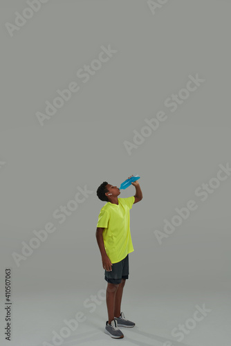 Refreshing after training. Vertical shot of a sporty teenage african boy holding blue energy drink while standing against grey background