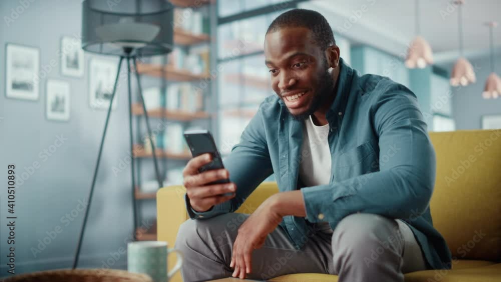 Excited Black African American Man Using Smartphone while Sitting on a Sofa in Living Room. Happy Man Smiling at Home and Chatting to Colleagues and Clients Over the Internet. Using Social Networks. Excited Black African American Man Using Smartphone while Sitting on a Sofa in Living Room. Happy Man Smiling at Home and Chatting to Colleagues and Clients Over the Internet. Using Social Networks.