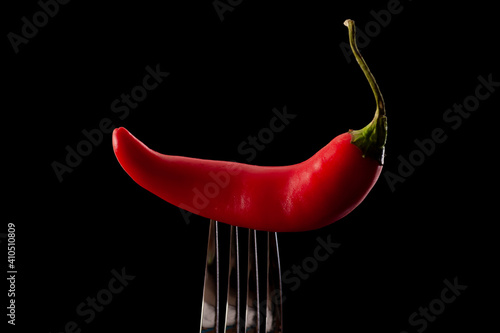 Mexican food, exotic taste and spicy condiments concept with red hot chili pepper stuck in a metal fork isolated on moody black background