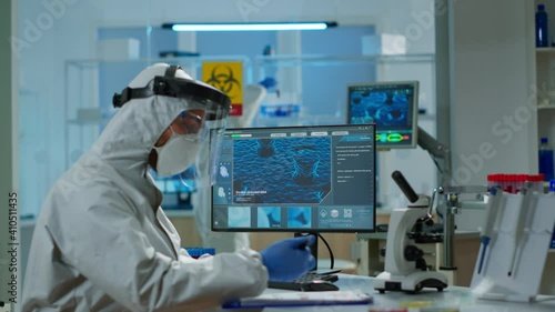 Scientist in protection suit typing on pc working in medical laboratory, writing on clipboard, developing inovative drugs against new virus. Doctor using high tech, chemistry tools for researching photo