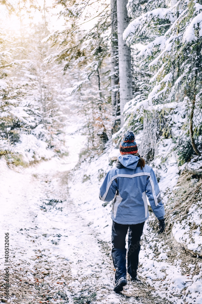 Young woman in a sports suit walks down the mountain path. Warm sunlight seeable on horizont. Hiker walking through the woods on snow trail path in winter. Cold weather hiking