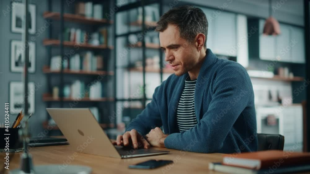 Handsome Caucasian Man Working on Laptop Computer while Sitting Behind Desk in Cozy Living Room. Freelancer Working From Home. Browsing Internet, Using Social Networks, Having Fun in Flat. Handsome Caucasian Man Working on Laptop Computer while Sitting Behind Desk in Cozy Living Room. Freelancer Working From Home. Browsing Internet, Using Social Networks, Having Fun in Flat.