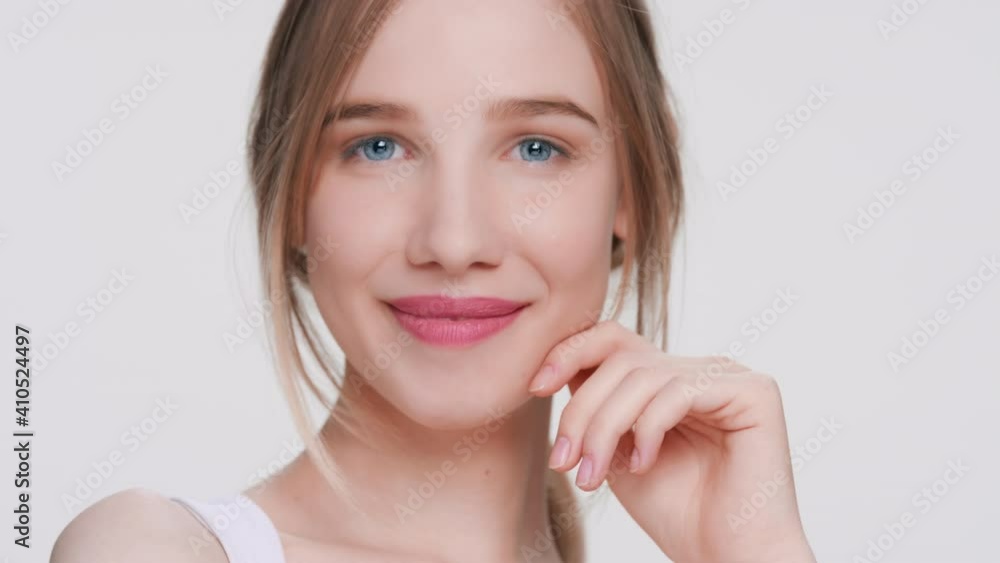 Close-up beauty portrait of young woman with blue eyes gently touches her face, runs finger from her cheek down to the chin looks at the camera and smiles | beauty products commercial concept Close-up beauty portrait of young woman with blue eyes gently touches her face, runs finger from her cheek down to the chin looks at the camera and smiles | beauty products commercial concept