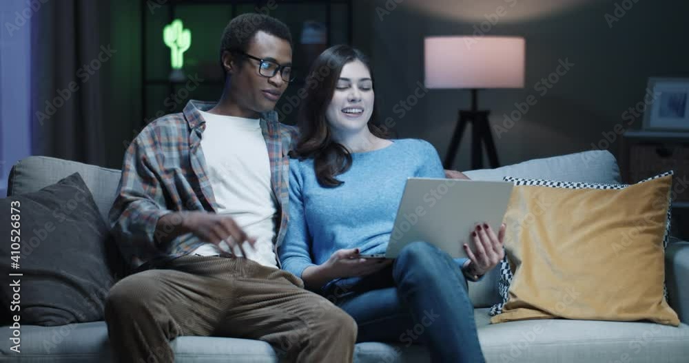 Young happy multinational couple sitting at night on couch at home and talking to friends on video call using laptop and waving hands. Young happy multinational couple sitting at night on couch at home and talking to friends on video call using laptop and waving hands.