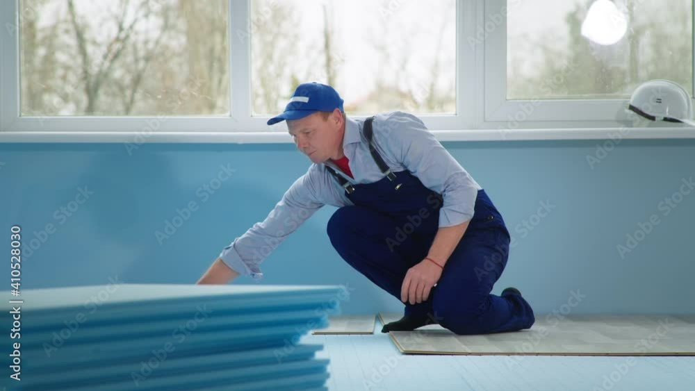 repair in new apartment, a professional man in work suit and cap on the floor lays the laminate on a blue substrate in the room near window repair in new apartment, a professional man in work suit and cap on the floor lays the laminate on a blue substrate in the room near window