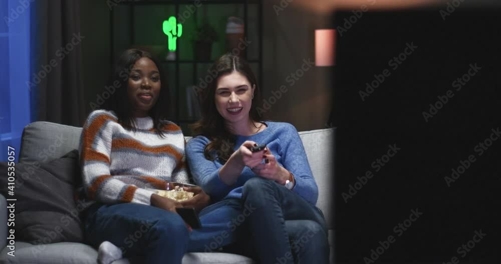 Two best friends sit on couch in evening and have fun watching comedy on TV and eating popcorn. Girlfriends gossip after reading message on phone. Two best friends sit on couch in evening and have fun watching comedy on TV and eating popcorn. Girlfriends gossip after reading message on phone.
