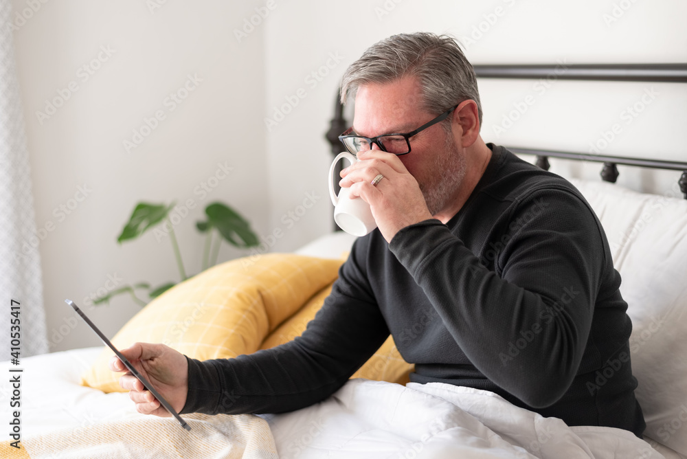 Fototapeta premium Man drinking coffee and reading a tablet in bed