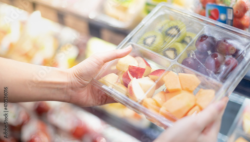 Female hand holding mix of fresh fruit in the transparent plastic box with fork in the grocery supermarket.