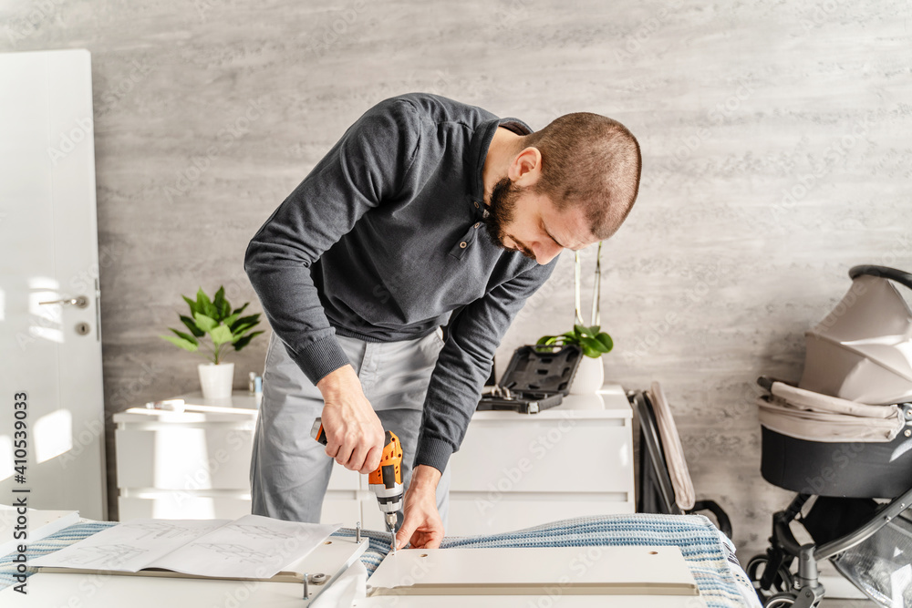 One Man with beard alone Putting Together Self Assembly Furniture at Home holding electric screwdriver looking the instructions - half length front view DIY concept real people copy space