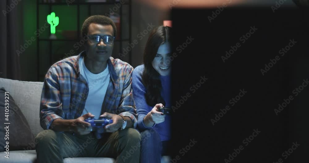 Young multinational couple playing video game on console while sitting at home in evening. Two people have fun jokingly pushing. Young multinational couple playing video game on console while sitting at home in evening. Two people have fun jokingly pushing.