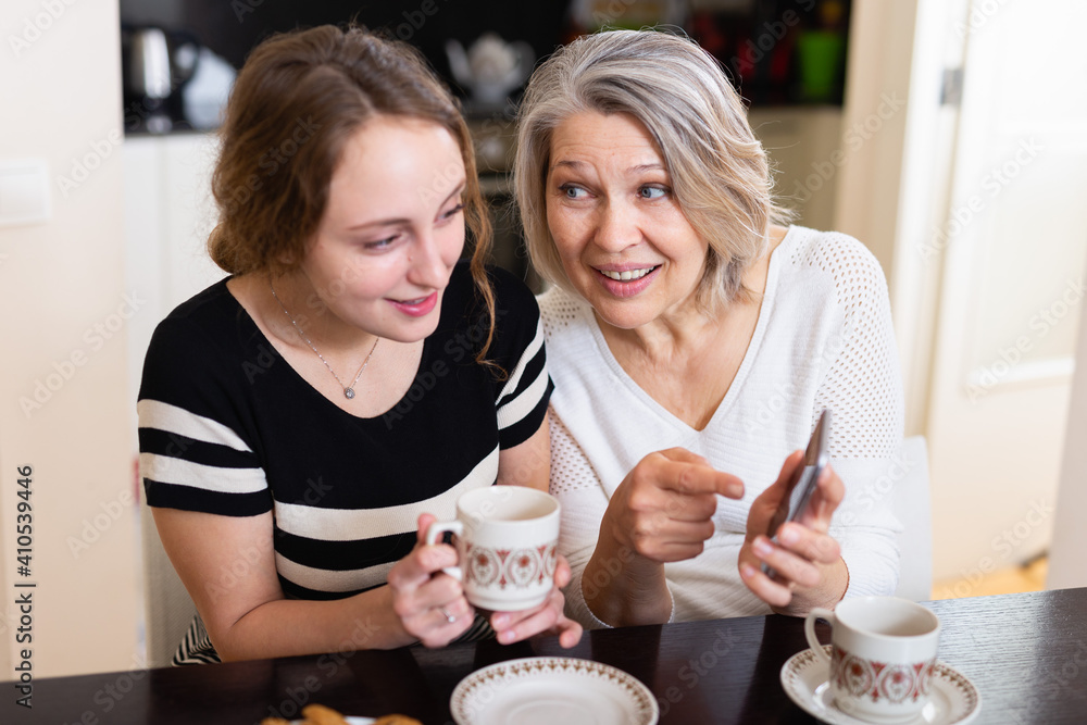 Happy mother and daughter using mobile gadget at kitchen table at home